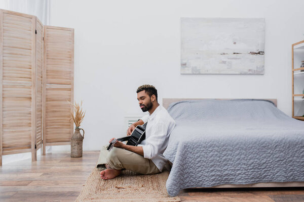 молодой африканский американец с окрашенными волосами сидит на ковре и играет на акустической гитаре возле кровати