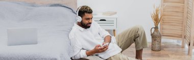 Yatakta dizüstü bilgisayarın yanında otururken elinde akıllı telefon tutan kablosuz kulaklıklı genç Afro-Amerikan bir adam.