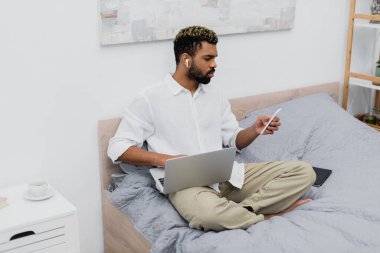 Genç Afro-Amerikalı kablosuz kulaklıklı adam yatakta otururken dizüstü bilgisayar ve akıllı telefon kullanıyor.