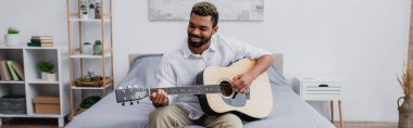 Memnun Afro-Amerikan adam boyalı saçlı ve sakallı yatak odasında akustik gitar çalıyor.