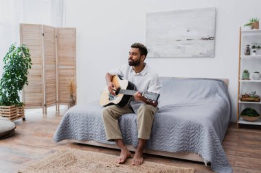 Çıplak ayaklı Afro-Amerikalı boyalı saçlı ve sakallı bir adam yatak odasında akustik gitar çalıyor.
