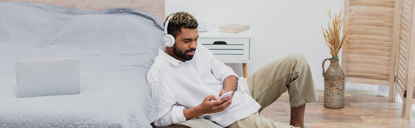 молодой африканский американец в беспроводных наушниках, держащий смартфон, сидя рядом с ноутбуком на кровати, баннер