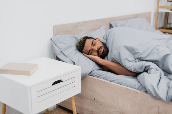 bearded african american man with closed eyes sleeping in bed near bedside table with book