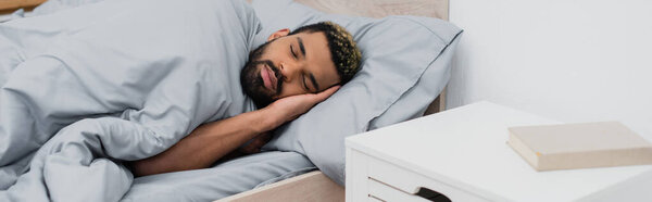 bearded african american man with closed eyes sleeping in bed near bedside table with book, banner