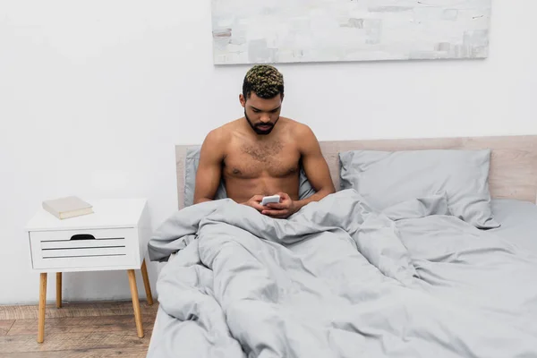 Shirtless Αφροαμερικανός Άνδρας Βαμμένα Μαλλιά Κουβεντιάζοντας Στο Smartphone Στο Υπνοδωμάτιο — Φωτογραφία Αρχείου
