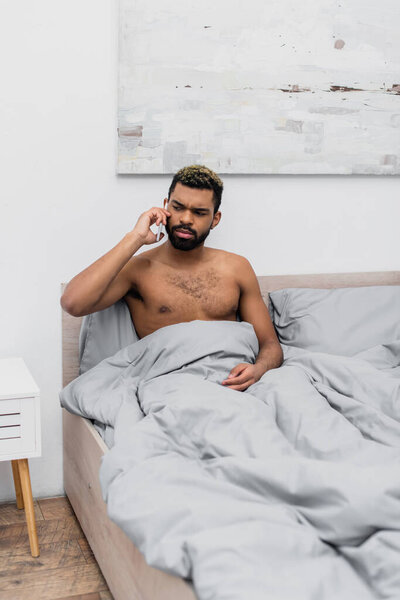 shirtless african american man with dyed hair talking on mobile phone in bed