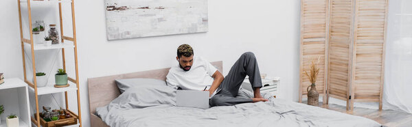бородатый африканский американец в беспроводном наушнике с помощью ноутбука и работает из дома в постели, баннер