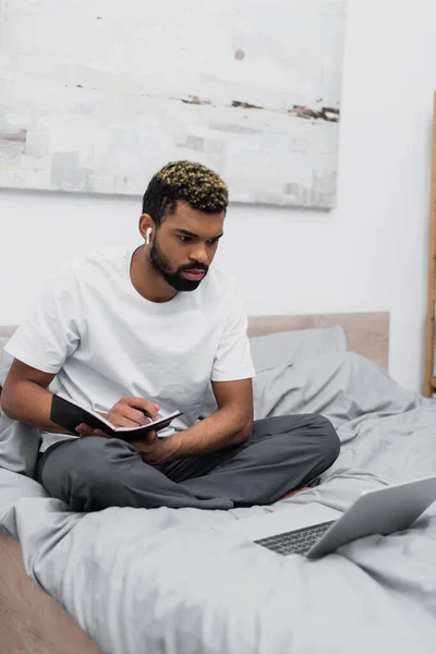 Зосереджений Афроамериканський Чоловік Бездротових Навушниках Використовуючи Ноутбук Тримаючи Блокнот Ліжку — стокове фото