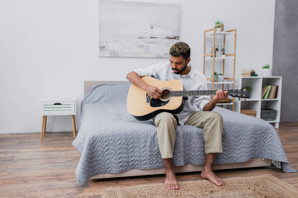 barefoot young african american man with dyed hair and beard playing acoustic guitar in bedroom
