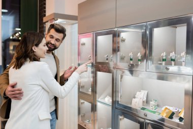 Man hugging girlfriend pointing with finger at jewelry in shop 