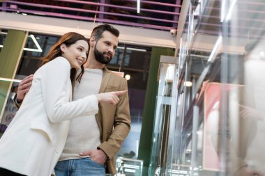 Low angle view of man hugging smiling girlfriend near showcase of jewelry shop 