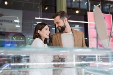 Smiling customer pointing at glass showcase near girlfriend in jewelry store 