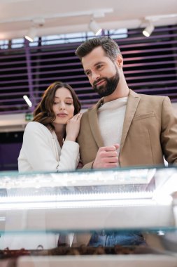 Brunette woman hugging boyfriend near blurred jewelry in shop 