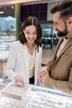 Smiling woman choosing rings near blurred boyfriend in jewelry store 
