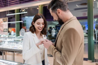 Smiling man wearing ring on hand of girlfriend in jewelry shop 