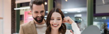Smiling woman holding earring near mirror and boyfriend in jewelry store, banner 