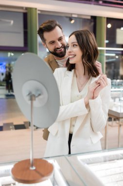 Cheerful man looking at girlfriend near blurred mirror in jewelry store 