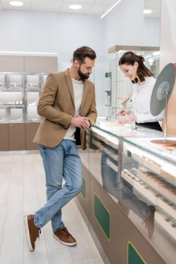 Smiling seller pointing at jewelry near customer in store 