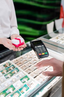 Cropped view of seller holding gift box near showcase and customer in jewelry shop 