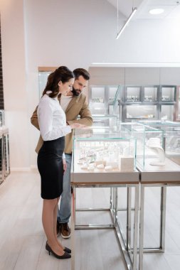 Man choosing jewelry accessories near seller pointing at showcase in shop 