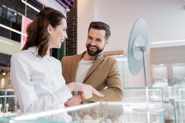 Positive customer looking at young seller pointing at showcase in jewelry shop