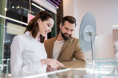 Brunette seller pointing at glass showcase near mirror and blurred client in jewelry shop 