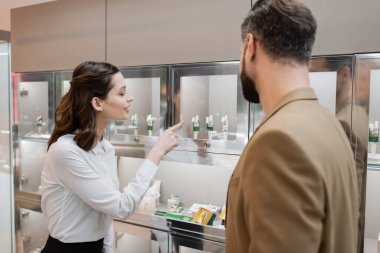 Smiling seller pointing at jewelry accessories near customer in store  