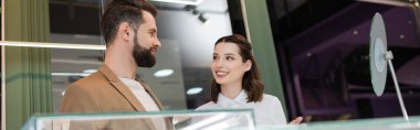 Positive seller talking to bearded customer near blurred showcase in jewelry store, banner 