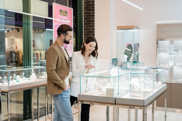 Smiling young woman pointing at jewelry near bearded boyfriend in store 