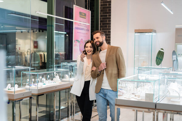 Cheerful young woman pointing at showcase while walking with boyfriend in jewelry shop 
