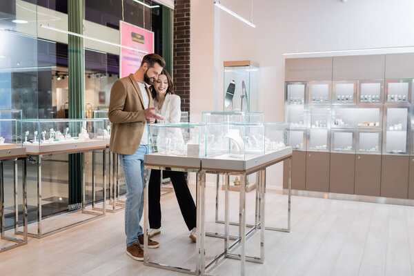 Bearded man pointing with hand at jewelry near brunette girlfriend in store 