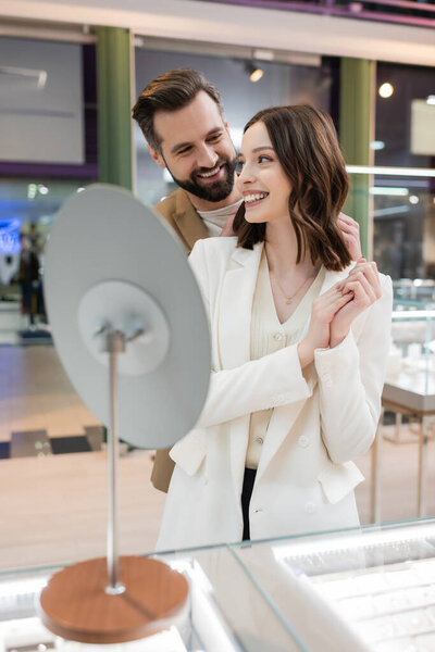 Cheerful man looking at girlfriend near blurred mirror in jewelry store 