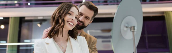 Smiling man wearing necklace on girlfriend in jewelry store, banner