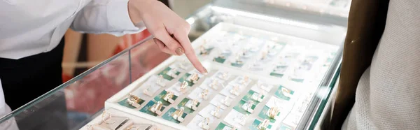 Cropped view of seller pointing at jewelry rings in glass showcase near customer in shop, banner