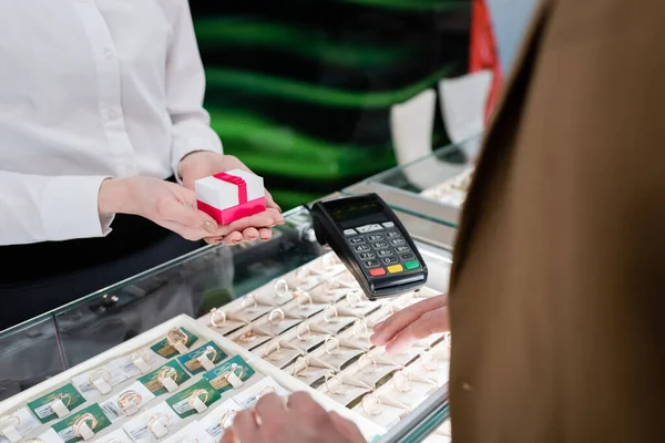 Cropped view of seller holding gift box near customer and credit card reader in jewelry store