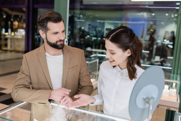 Customer looking at smiling seller pointing at glass showcase in jewelry shop 