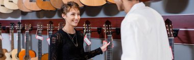 Smiling buyer holding acoustic guitar near african american seller in music shop, banner 