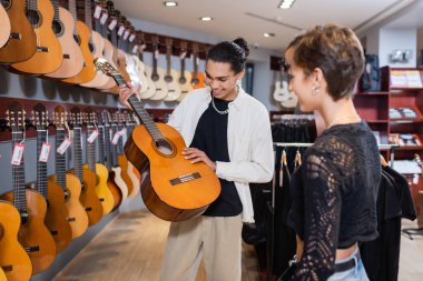Cheerful african american seller holding acoustic guitar near blurred woman in music store
