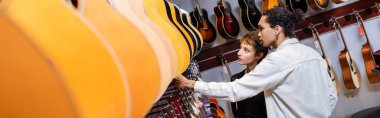 African american seller and customer looking at blurred acoustic guitars in music store, banner 