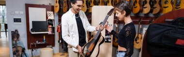 KYIV, UKRAINE - FEBRUARY 16, 2022: Smiling buyer looking at electric guitar near african american seller in music shop, banner 