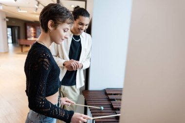 Smiling customer playing xylophone near blurred african american seller in music store clipart