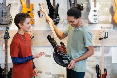 KYIV, UKRAINE - FEBRUARY 16, 2022: Smiling african american customer holding electric guitar near seller pointing with hand in music store 