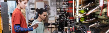 Side view of seller using digital tablet near african american customer and electronic drums in music shop, banner 
