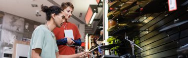 African american customer playing electronic drums near seller with digital tablet in music store, banner 
