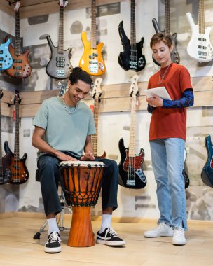 KYIV, UKRAINE - FEBRUARY 16, 2022: Positive african american customer playing djembe near seller with digital tablet in music store 
