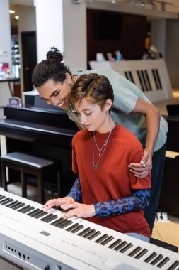 Cheerful african american man hugging girlfriend playing synthesizer in music shop 