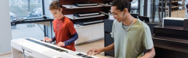 Smiling african american musician playing synthesizer near girlfriend in music shop, banner 