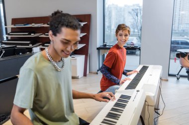 Cheerful woman playing synthesizer and looking at blurred african american boyfriend in music shop 