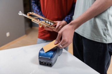 Cropped view of african american man paying with credit card near blurred girlfriend with trumpet in music store 