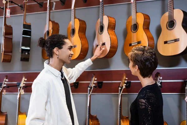 Smiling african american seller pointing at acoustic guitar near buyer in music store